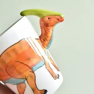 Weiße Keramiktasse mit orangem Dinosaurier-Motiv und grünem Dinosaurier-Kopf als Henkel